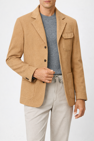 Man wearing a tan blazer over a gray shirt and light-colored pants on a white background